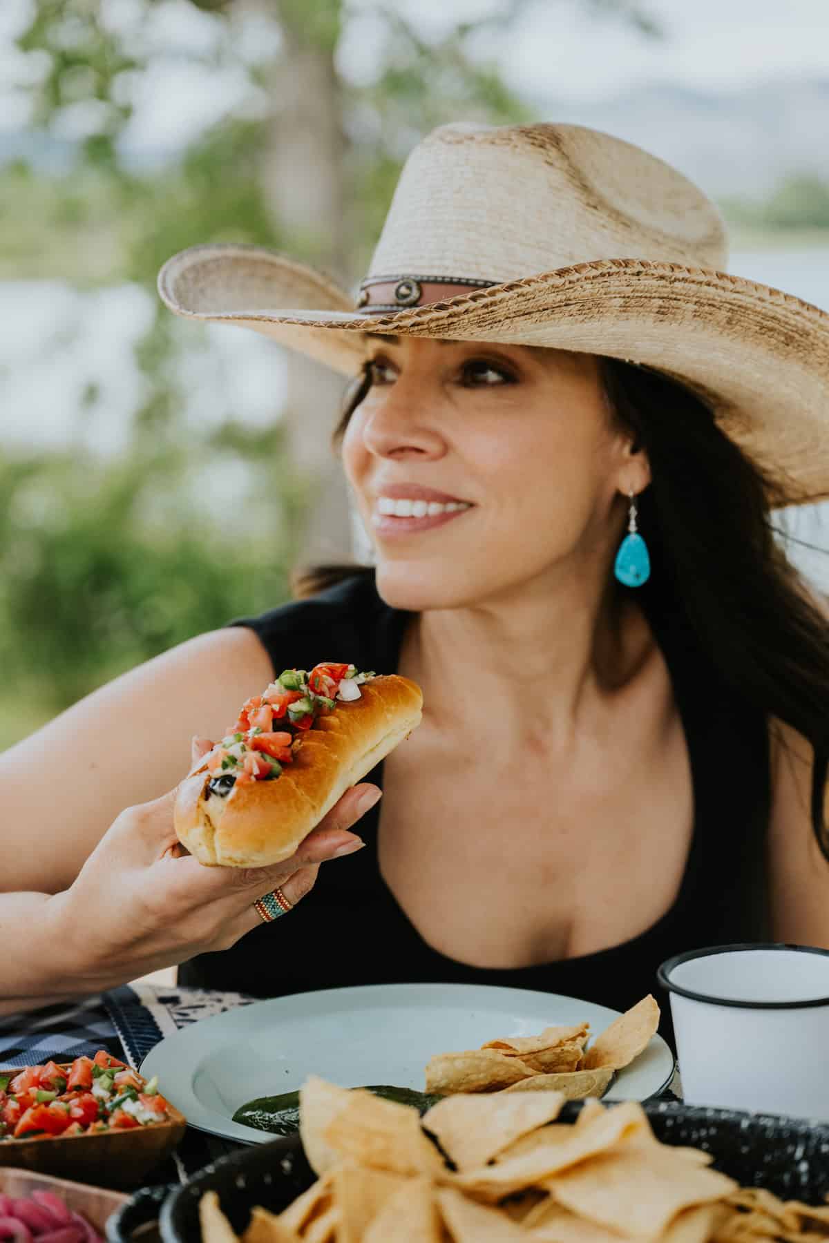 Yvette Marquez, author of Muy Bueno, wearing a cowboy hat, turquoise drop earrings, and a black tank top holding a Mexican hot dog with bacon, pico de gallo, and guacamole at a picnic table. 