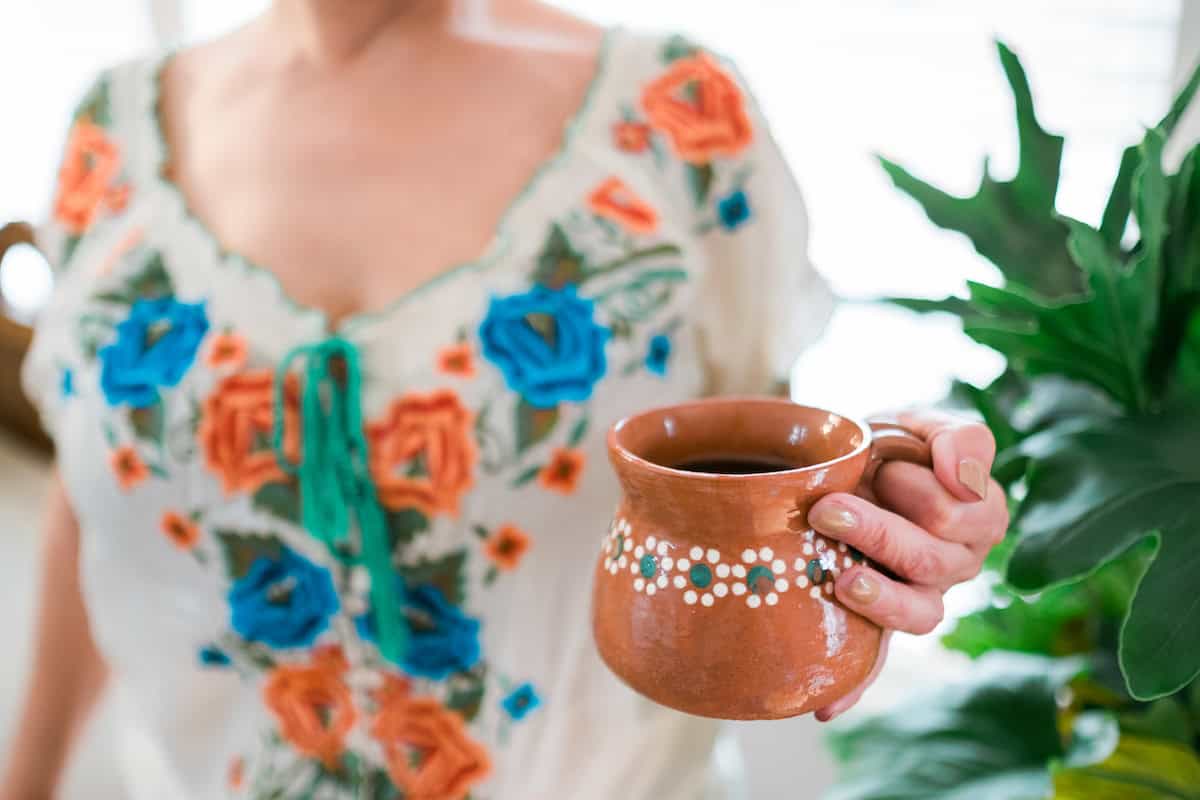 woman in a pretty turquoise, aqua, orange, and white embroidered floral top holding a terracotta mug of Mexican té de canela. 