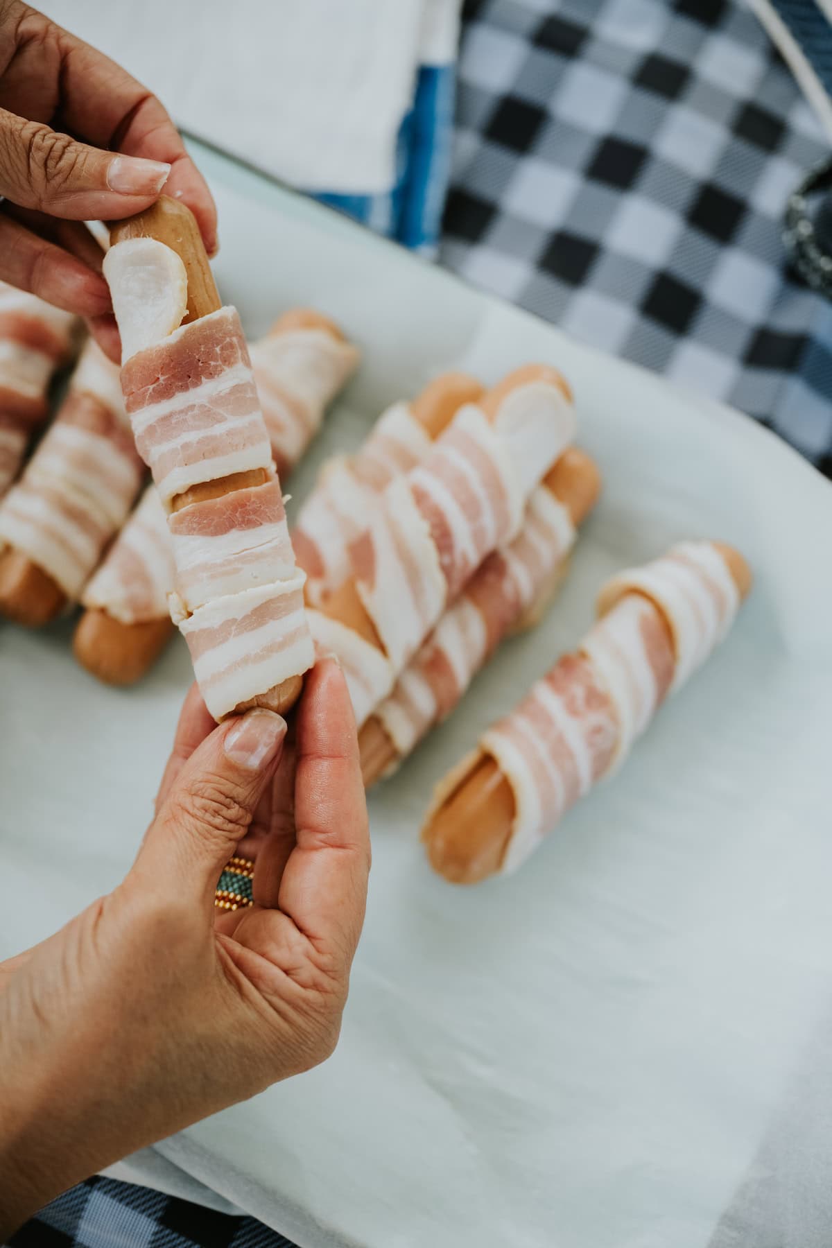 hand holding raw hot dog that has been wrapped in bacon with a platter of more raw bacon wrapped hot dogs below. 