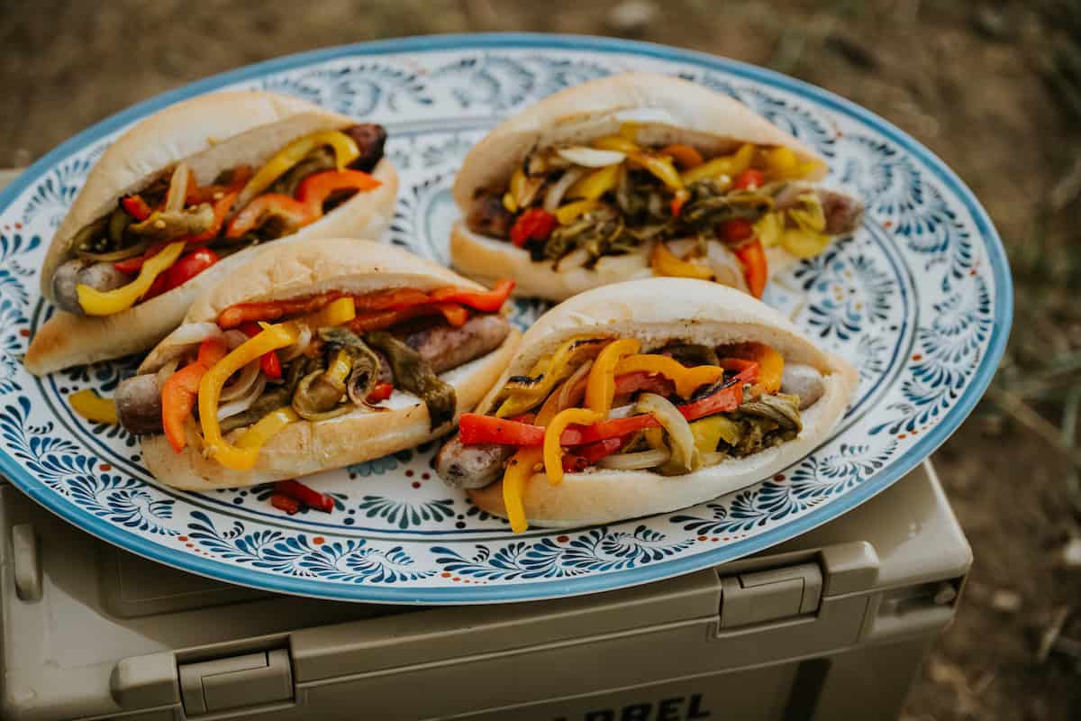 4 assembled grilled beer brats with peppers, onions, and roasted chiles on crusty bolillo rolls on a blue and white oval serving platter.