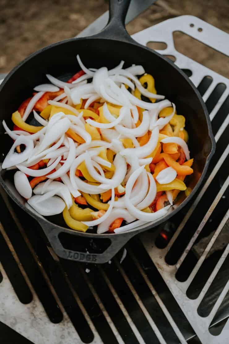 cast iron skillet on a grill with raw slices of multicolored bell peppers and white onions. 