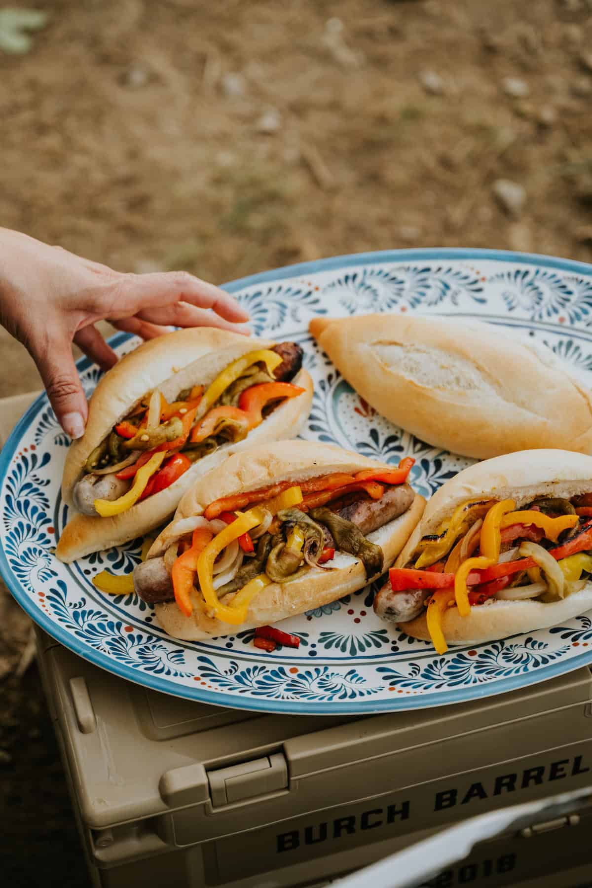 hand grabbing a grilled beer brat-stuffed bolillo roll topped with bell peppers, onions, and roasted hatch chiles off an oval blue and white serving platter of several that have been assembled. 