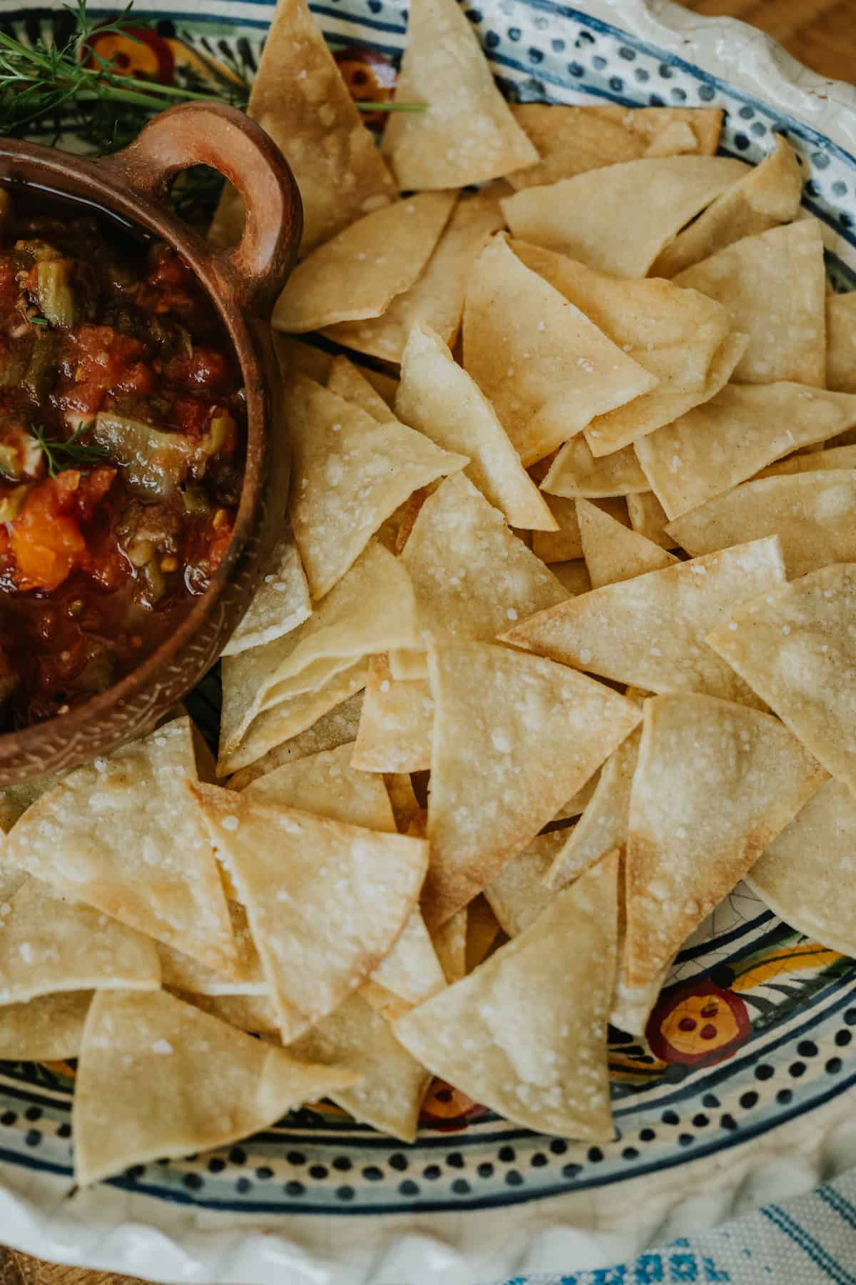 overhead shot of homemade corn tortilla chips on an oblong, handpainted serving platter with a rustic terracotta bowl filled with salsa casera. 