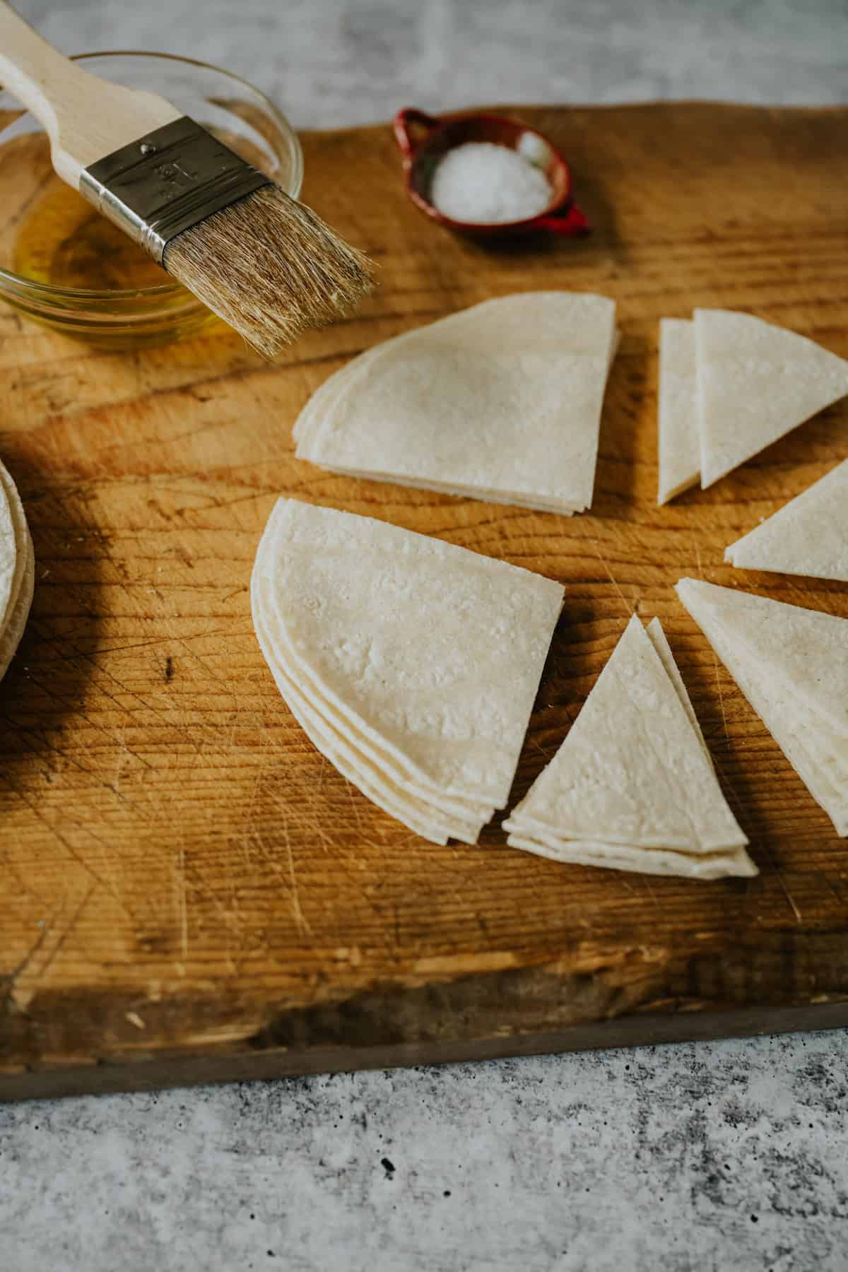 wooden cutting board with a stack of corn tortillas being cut into 8 wedges with a small bowl of oil, a pastry brush, and a small bowl of salt. 