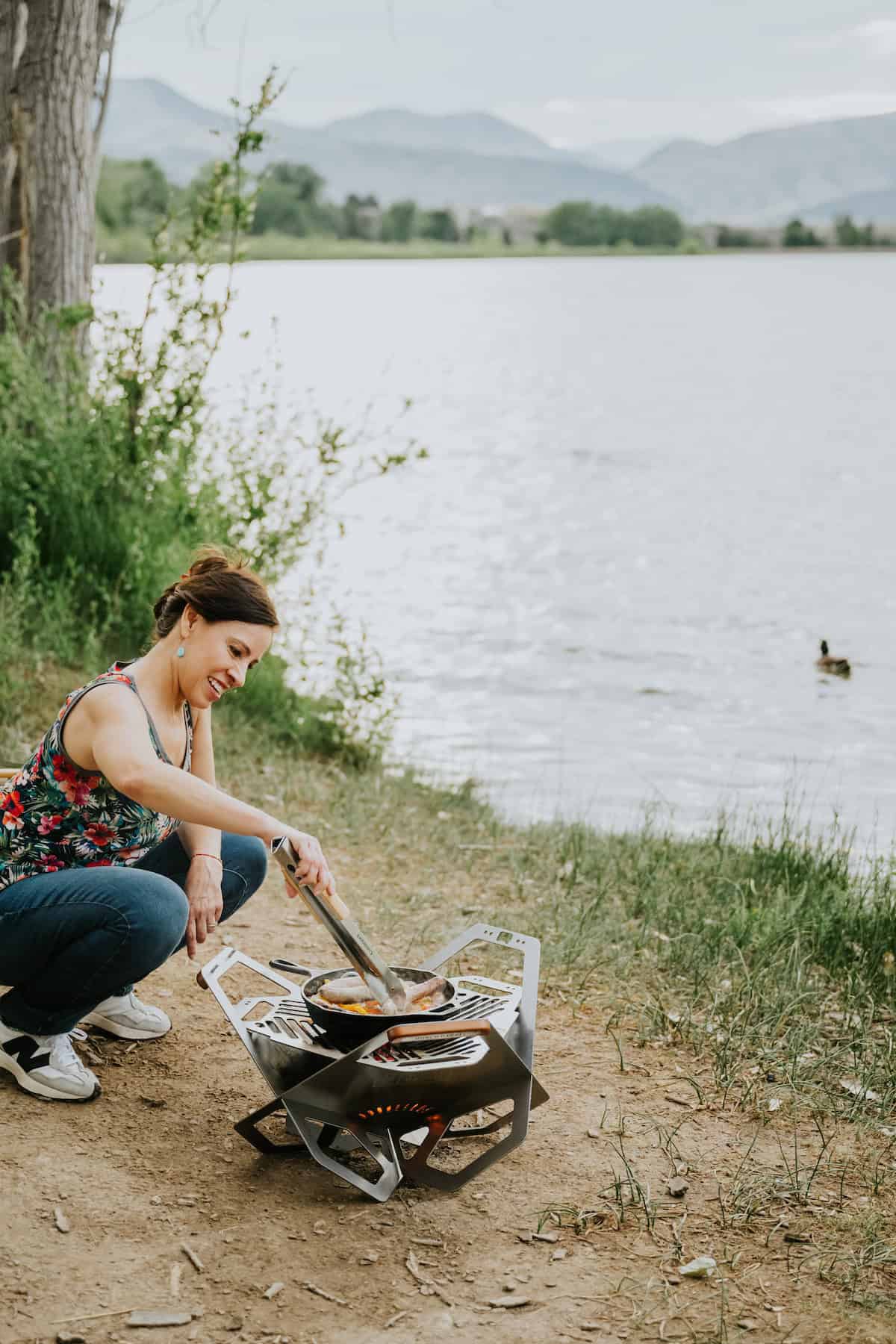 Blogger Yvette Marquez using a cast iron skillet on a camping stove to saute fajita veggies and grill beer brats in front of a lake. 