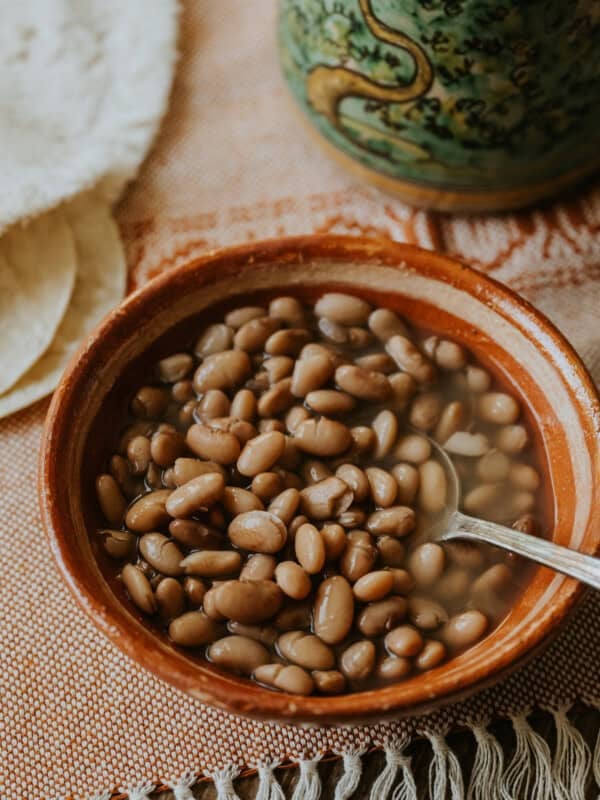 hand thrown terracotta bowl of frijoles de la olla with a silver spoon atop an orange and white textile with tortillas and a painted turquoise vase blurred in the background.