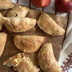 flat lay shot of a bunch of baked apple empanadas on a dark wooden tray with one broken in half to expose the spiced apple filling.