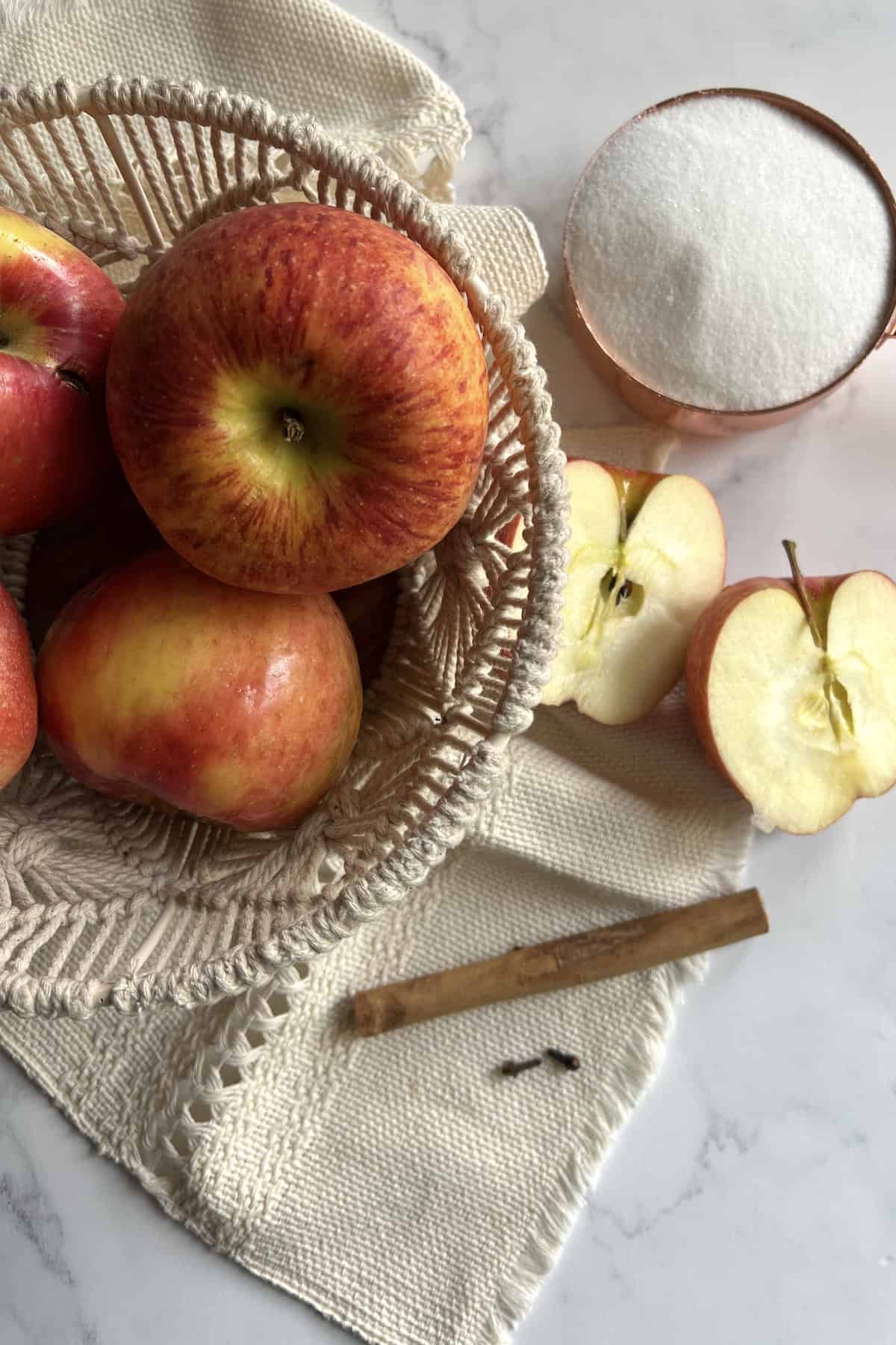 flat lay shot of a woven bowl filled with apples on a white table with a canela stick, two cloves, and a halved apple. 