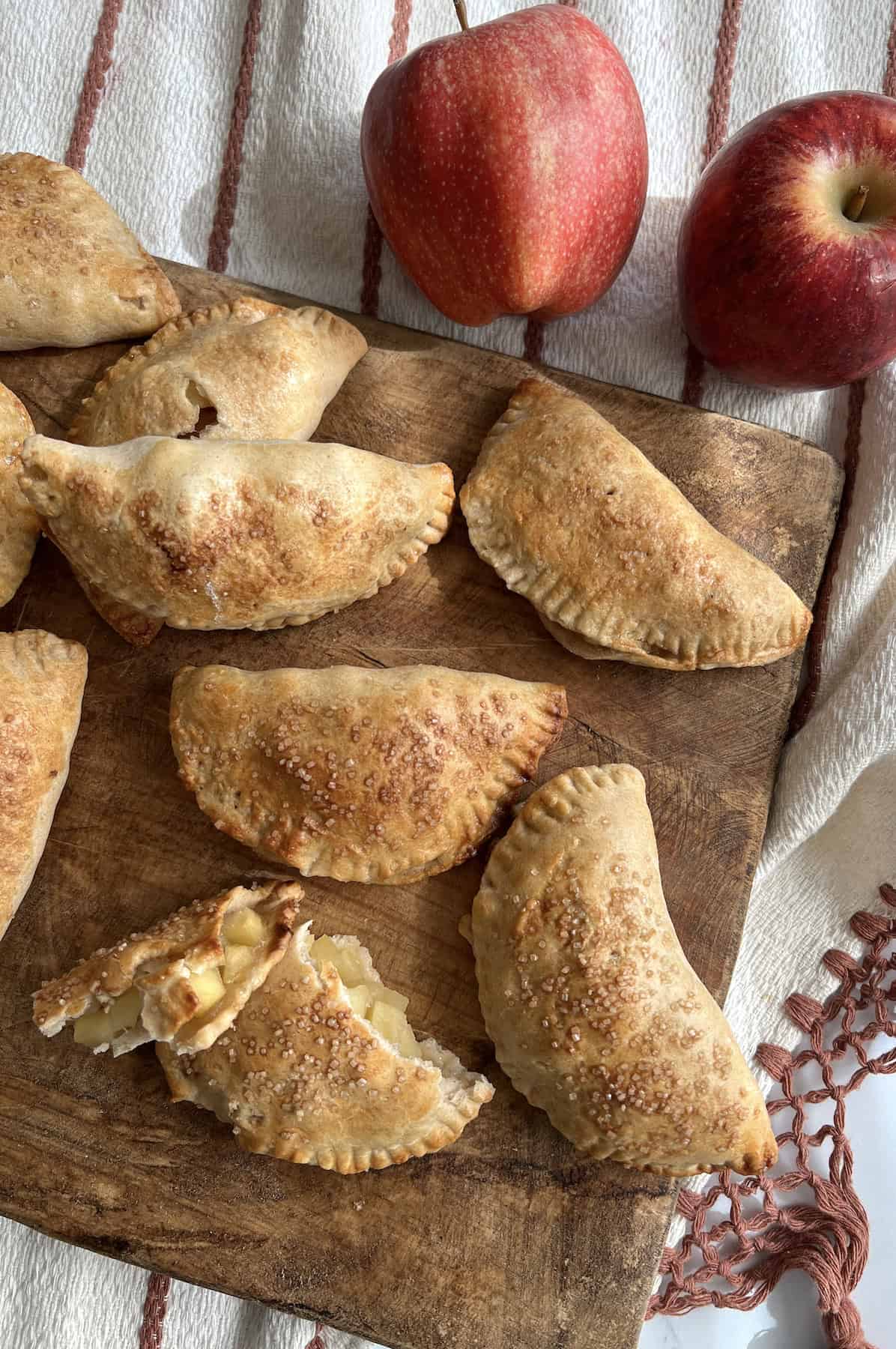 flat lay shot of a bunch of baked apple empanadas on a dark wooden tray with one broken in half to expose the spiced apple filling.