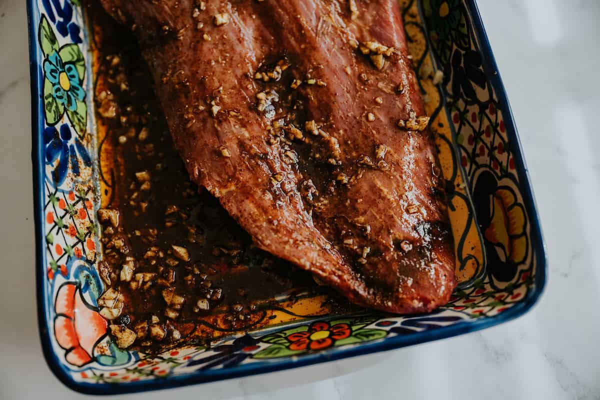 whole flank steak mixed with fajita marinade in a colorful hand-painted rectangular dish. 