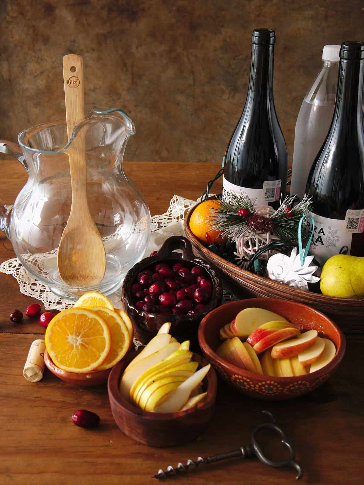 empty clear glass pitcher with a wooden spoon, 2 bottles of red wine, sliced oranges, pears, and apples, and whole cranberries in earthenware bowls in preparation for making christmas red sangria. 