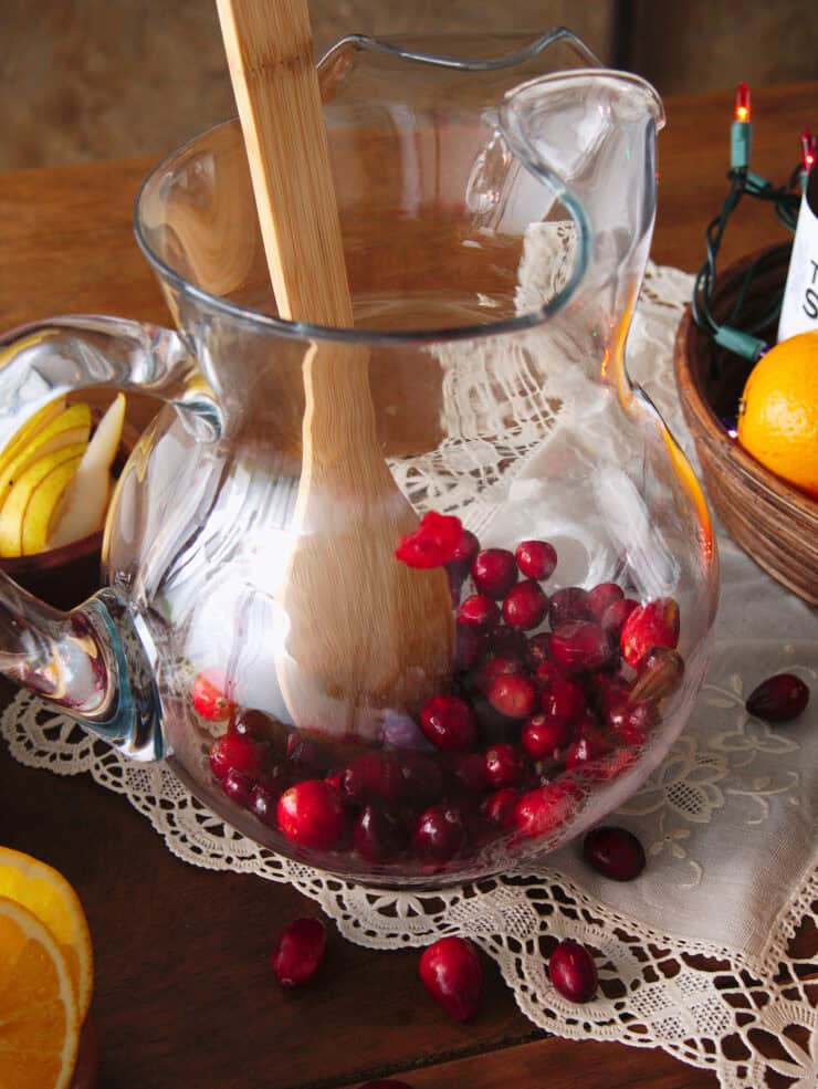 wooden kitchen spoon muddling cranberries in the base of a clear glass pitcher. 