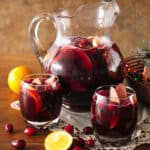 clear glass pitcher and two rocks glasses filled with fruit-filled red Christmas sangria on top of a white doily on a wooden table with cranberries scattered around and a bowl with multicolored christmas lights to the side.