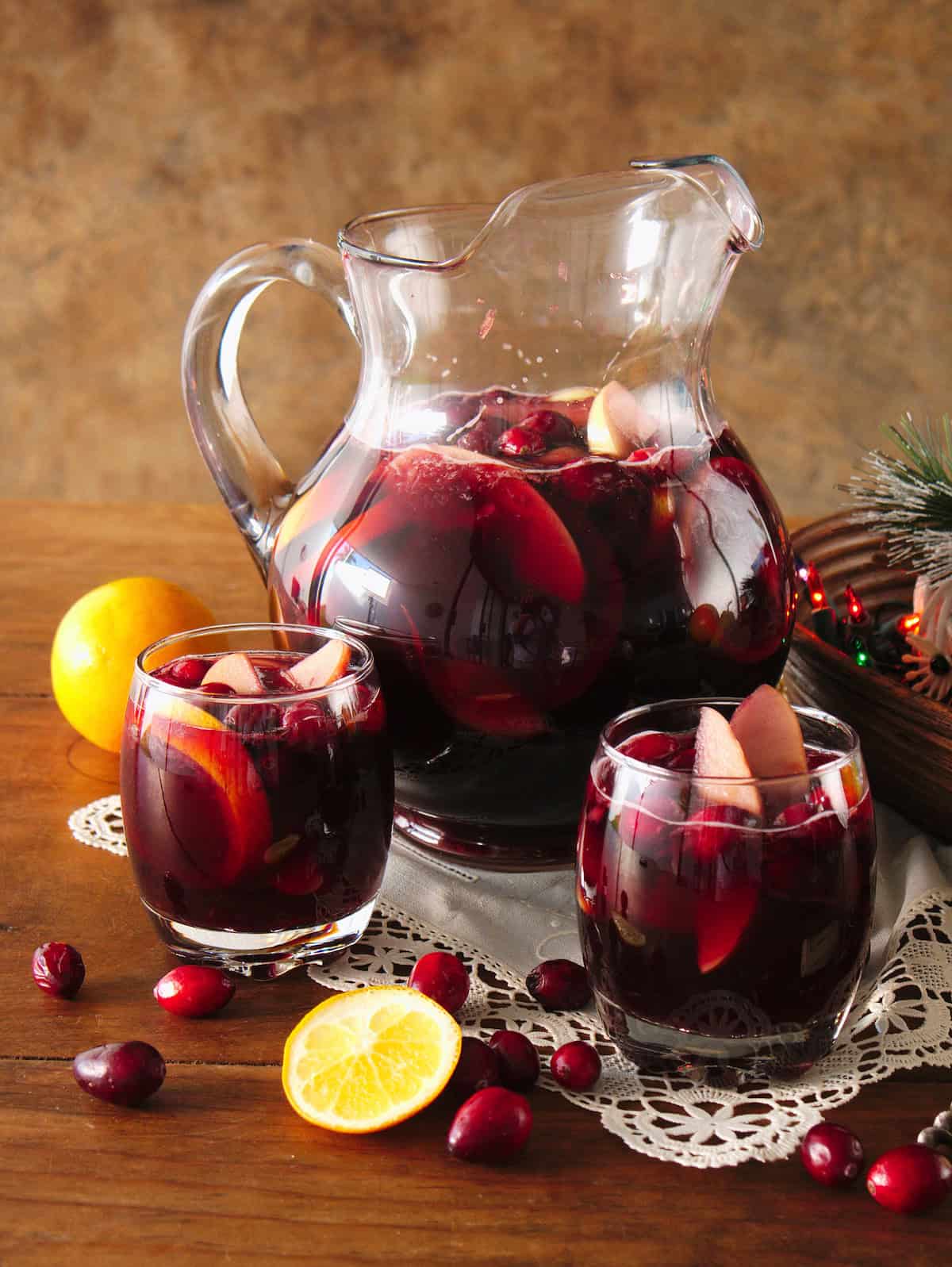 clear glass pitcher and two rocks glasses filled with fruit-filled red Christmas sangria on top of a white doily on a wooden table with cranberries scattered around and a bowl with multicolored christmas lights to the side. 