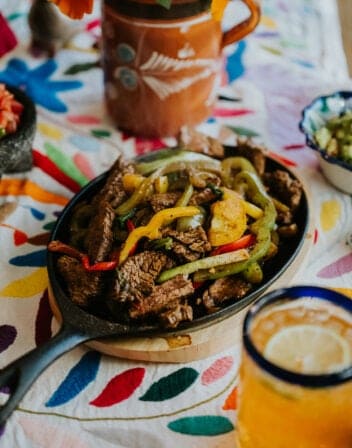 cast iron steak fajita skillet with peppers and green onions on a matching wooden tray on top of a colorful embroidered tablecloth with a vase of pink and orange flowers in the background and a blue-rimmed clear glass of beer in the foreground.