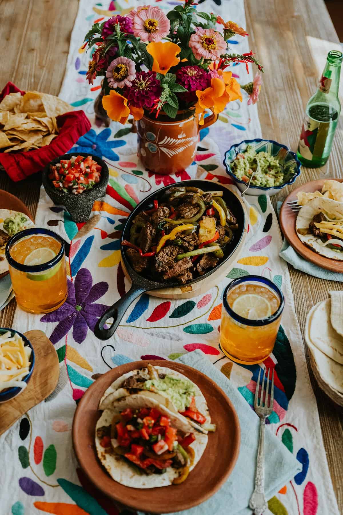 serving table with a colorful embroidered runner topped with a molcajete of fresh pico de gallo, a painted bowl filled with guacamole, a fajita serving skillet filled with steak fajitas and veggies, a basket of warm tortillas, and a bowl of shredded cheese with a few plates with assembled fajitas and some tortilla chips on the sides. 