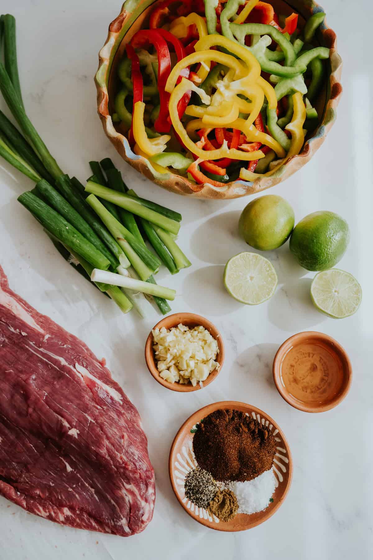 flat lay shot of a piece of flank steak, quartered green onions, minced garlic, spices, oil, halved limes, and sliced peppers and onions measured out for making steak fajitas. 