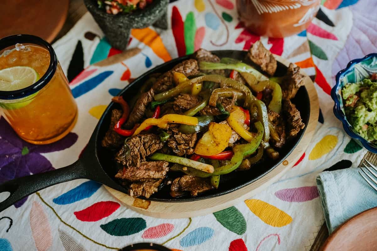 overhead shot of a flank steak fajitas skillet on a colorful embroidered table runner with a bowl of guacamole to the right, the corner of a molcajete above, and a glass of iced tea to the left. 