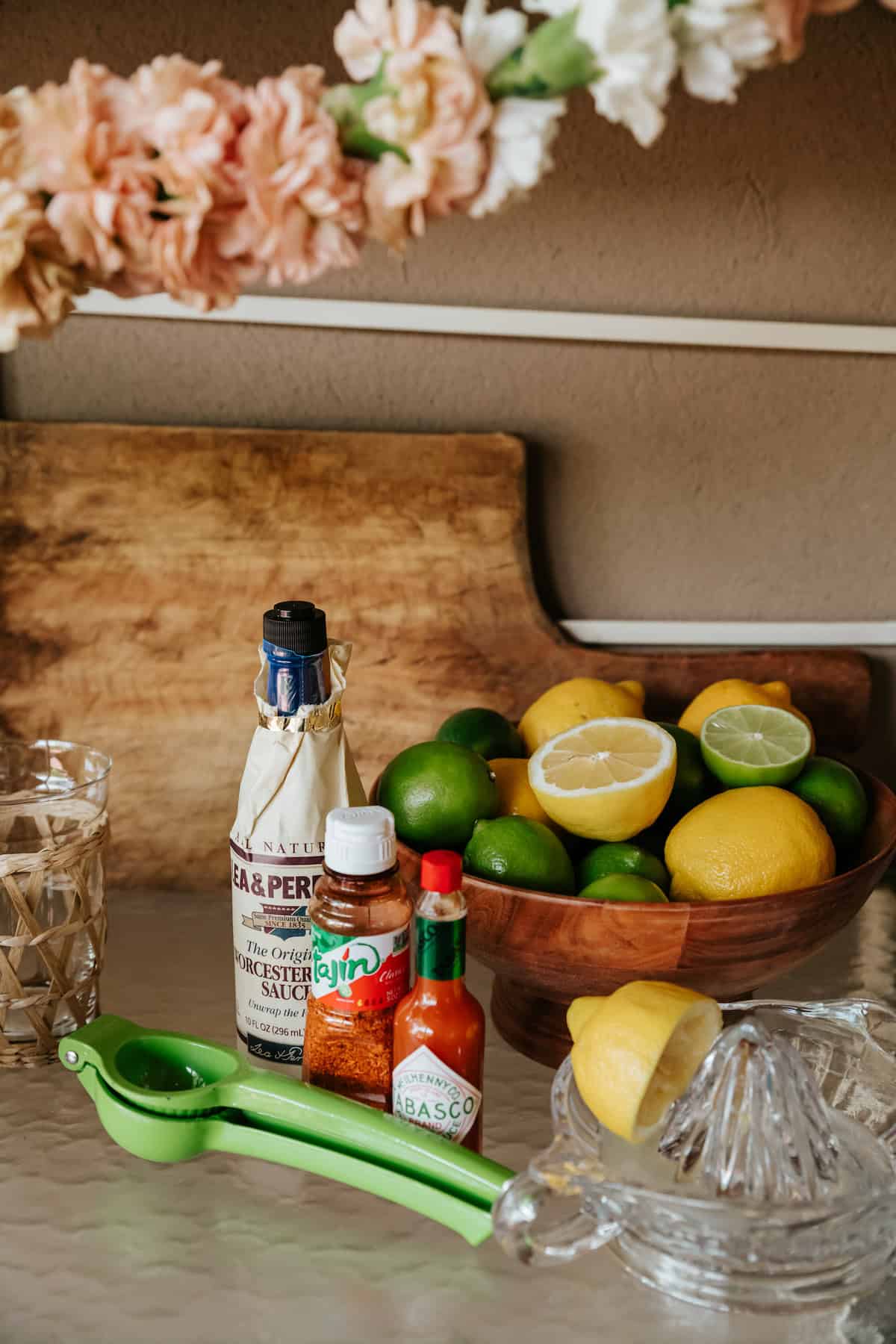 worcester sauce, tajin, and tabasco bottles on a glass table with a bowl of fresh lemons an limes in the background and a halved lemon on an old-fashioned citrus reamer and lime juice presser in the foreground.