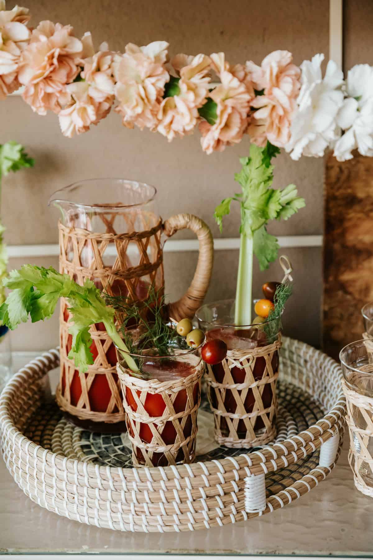 woven serving tray with a matching pitcher and two glasses with a woven rafia exterior filled with homemade bloody marias under a peach and white carnation garland.