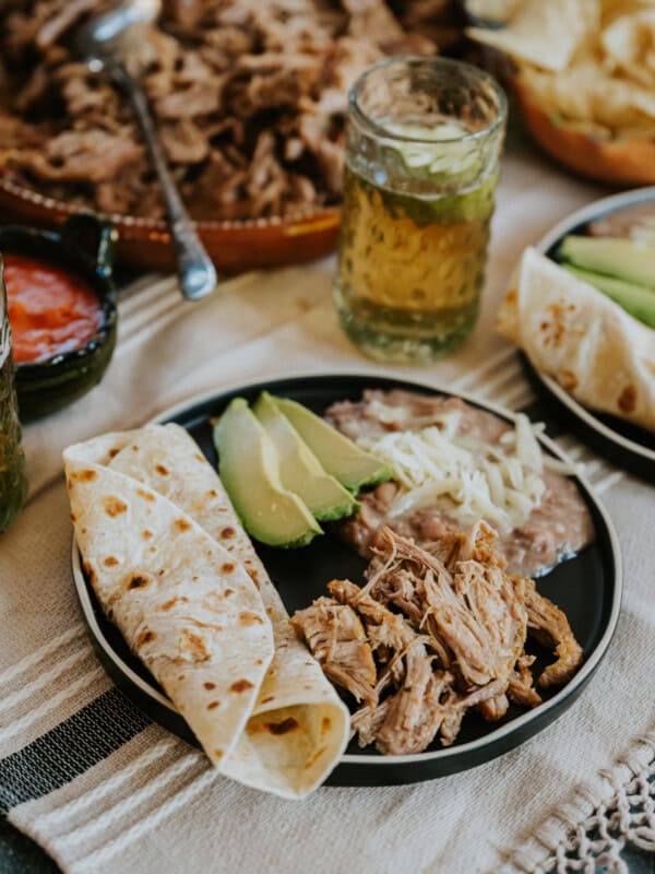 Dutch oven carnitas on a round black plate with a scoop of refried beans topped with cheese, slices of avocado, and a warmed tortilla on the side.