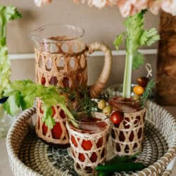 serving tray with a pitcher and two glass of spicy bloody marias garnished with celery, cherry tomatoes, olives, and dill with two serrano chiles in the foreground.