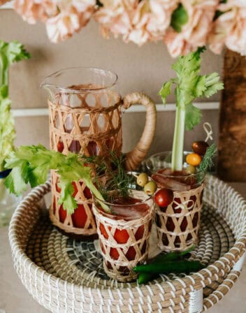 serving tray with a pitcher and two glass of spicy bloody marias garnished with celery, cherry tomatoes, olives, and dill with two serrano chiles in the foreground.