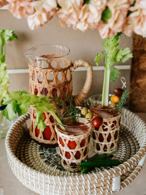 serving tray with a pitcher and two glass of spicy bloody marias garnished with celery, cherry tomatoes, olives, and dill with two serrano chiles in the foreground.