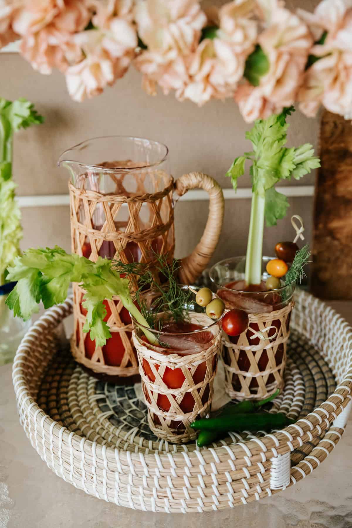 serving tray with a pitcher and two glass of spicy bloody marias garnished with celery, cherry tomatoes, olives, and dill with two serrano chiles in the foreground.