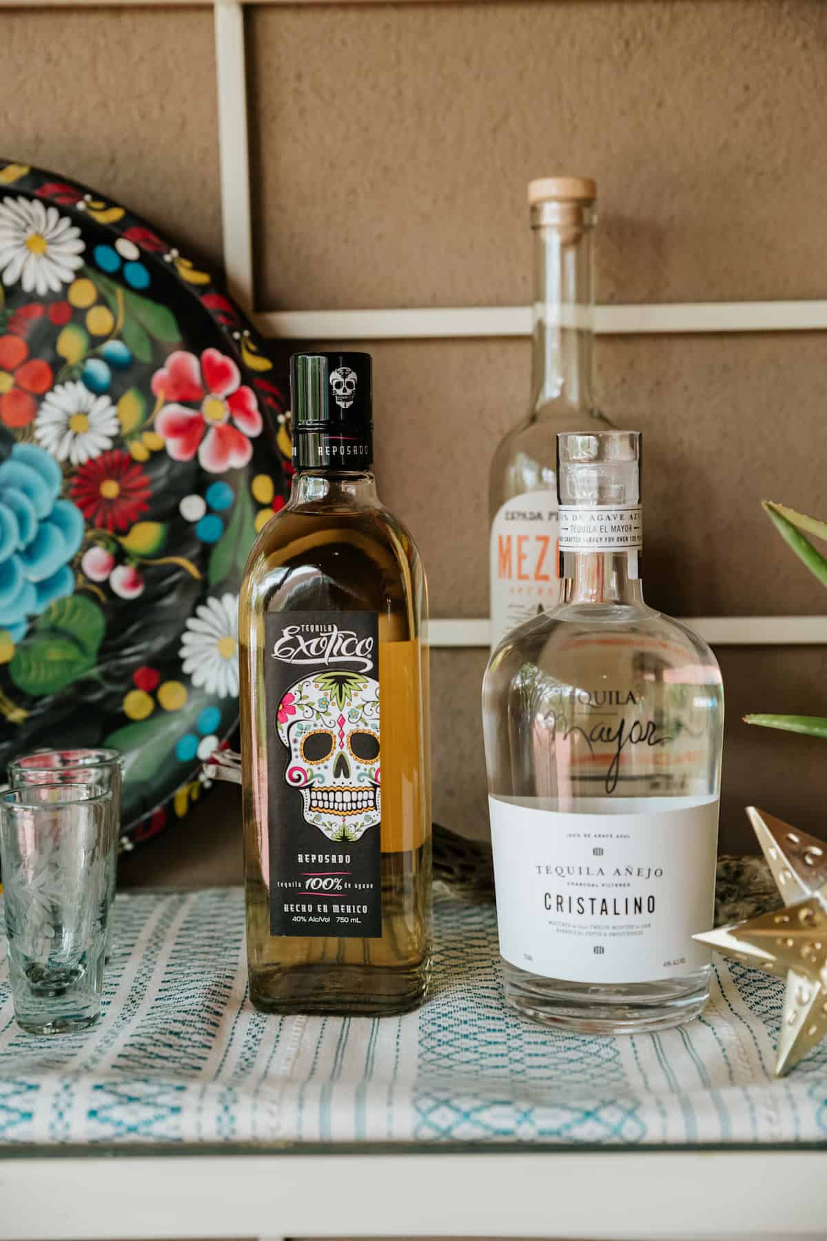 3 bottles of tequila—mezcal, cristalino, and reposado style—on a blue and white embroidered cloth with a colorful hand-painted black floral tray in the background.