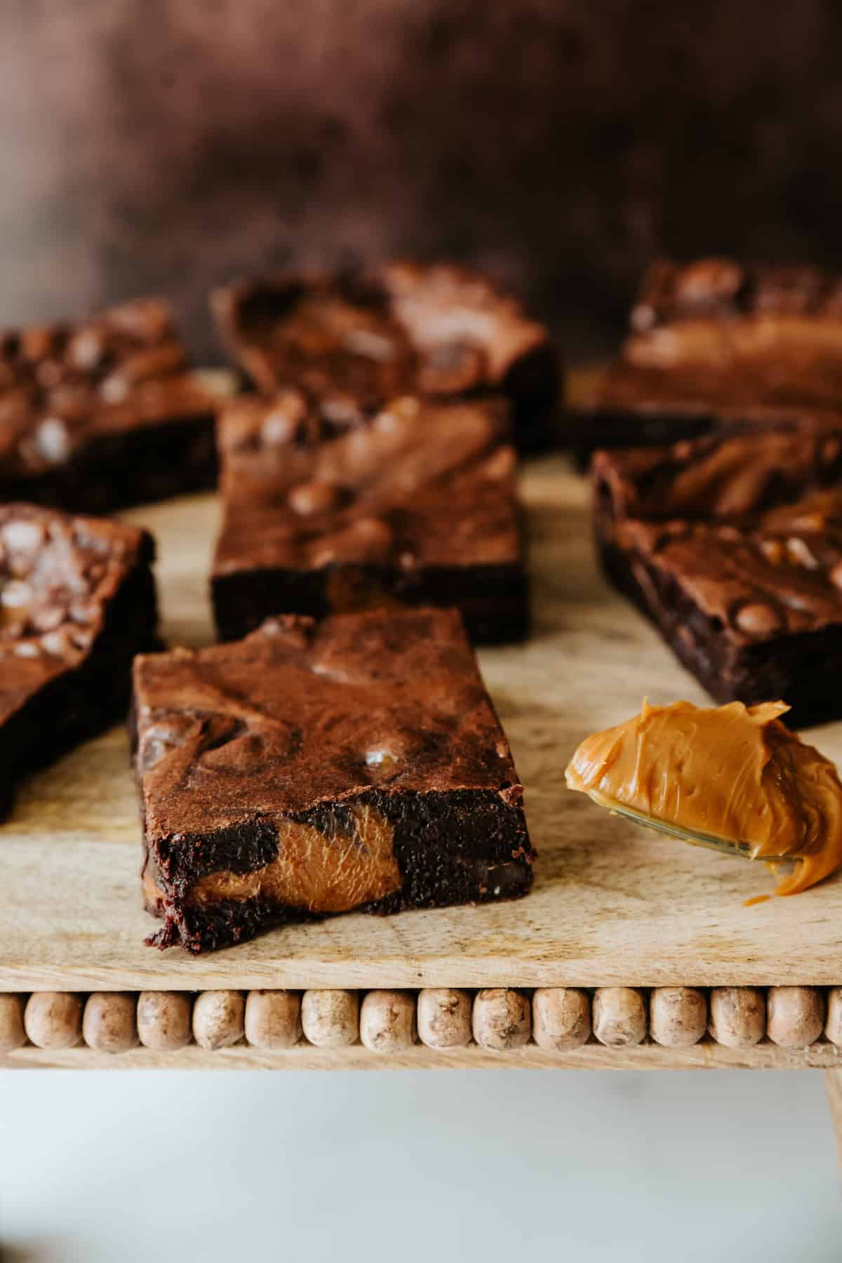 Dulce de leche brownie squares on a wooden pedestal with a brown background. 