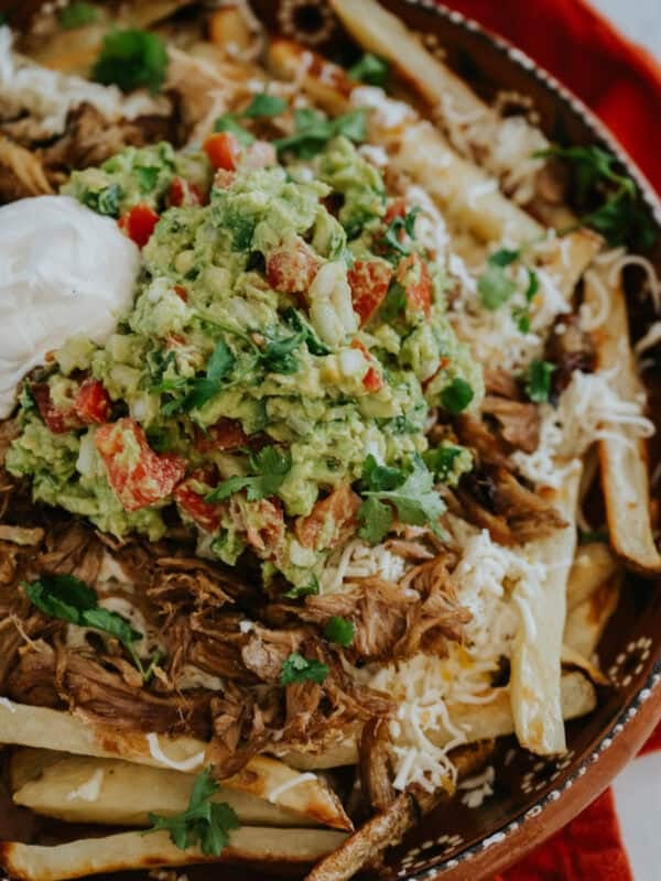 closeup shot of a pile of carnitas fries—oven baked french fries topped with cheese, carnitas, guacamole, and sour cream.