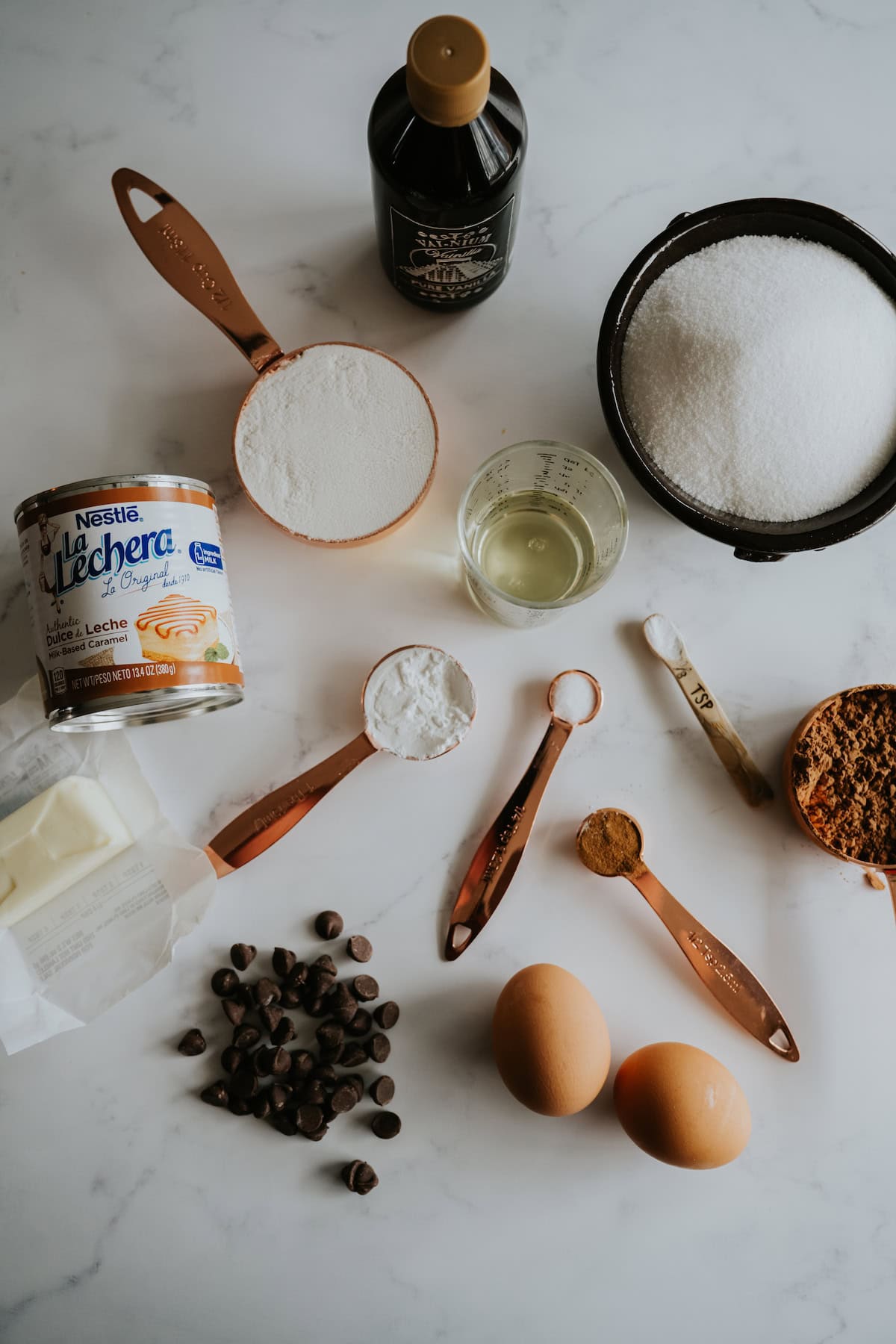 A variety of ingredients to make dulce de leche brownies. Vanilla extract in a bottle, dulce de leche in a can, wet and dry ingredients in measuring cups and spoons on a white marble background.