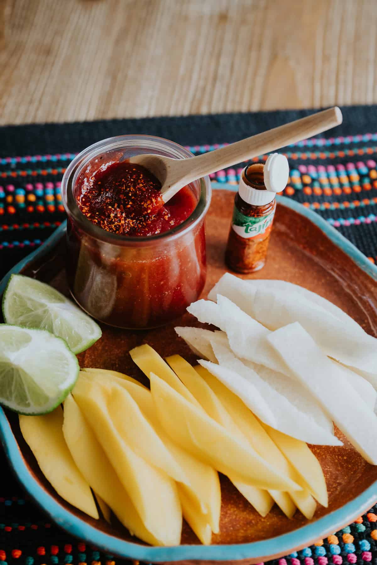 Homemade Mexican Chamoy Sauce in a preserve jar with a wooden spoon and sprinkled with chile lime seasoning and slices of mango and jicama on plate.