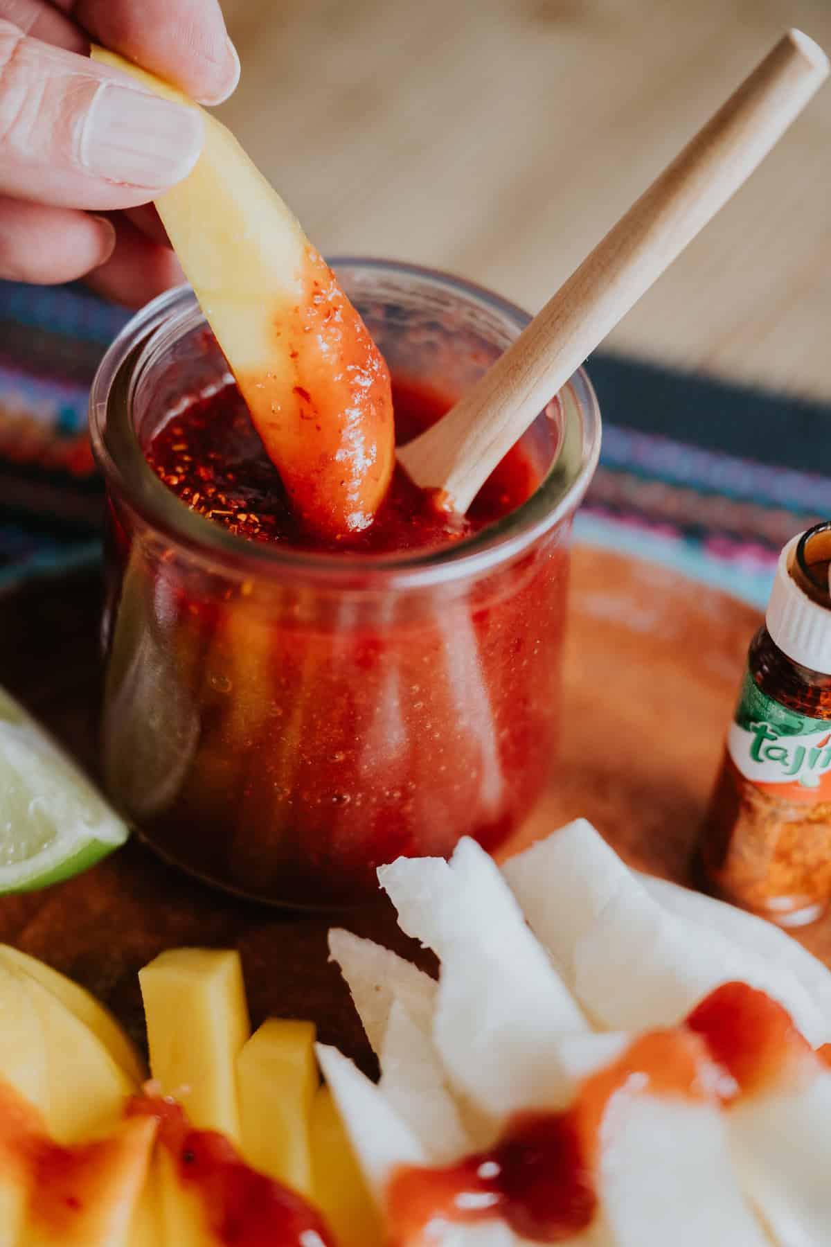 Slice on mango being dipped in homemade Mexican Chamoy Sauce in a clear jar with a wooden spoon and sprinkled with chile lime seasoning and slices of mango and jicama on plate.
