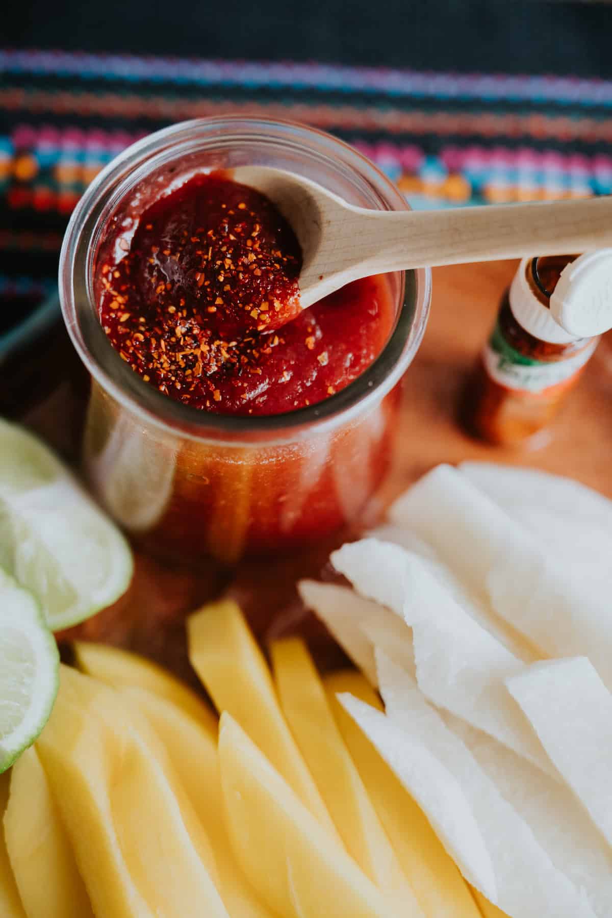 Homemade Mexican Chamoy Sauce in a preserve with a wooden spoon and sprinkled with chile lime seasoning and slices of mango and jicama on plate.