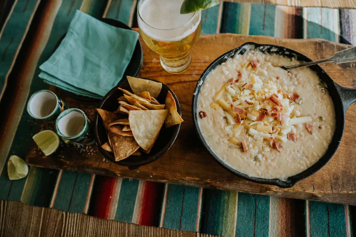 wooden table with a colorful striped Mexican blanket topped with a wooden tray with a light Mexican beer with a lim, a cast iron skillet with white refried bean and cheese dip topped with extra cheese and bacon bits, a black bowl of tortilla chips, and a stack of turquoise cocktail napkins. 