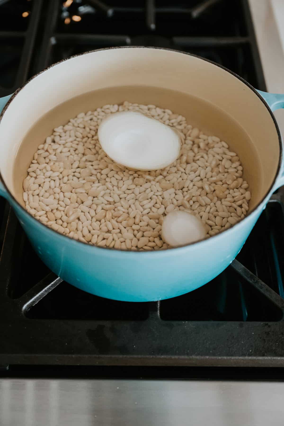 white beans in a turquoise enameled cast iron dutch oven with water and a quartered onion. 