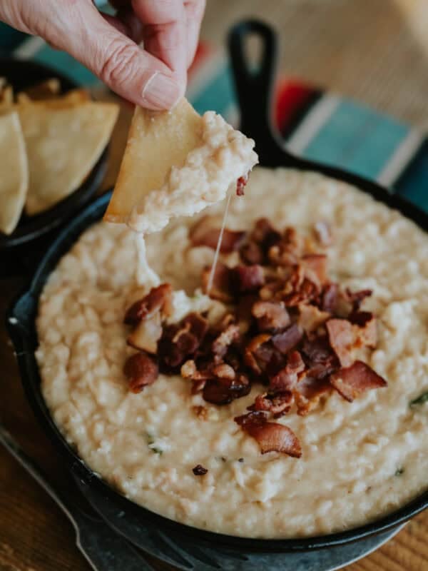 action shot of a tortilla chip being dipped into a bowl of cheesy white refried bean dip topped with extra bacon bits; the cheese is stretching from the bowl to the chip.