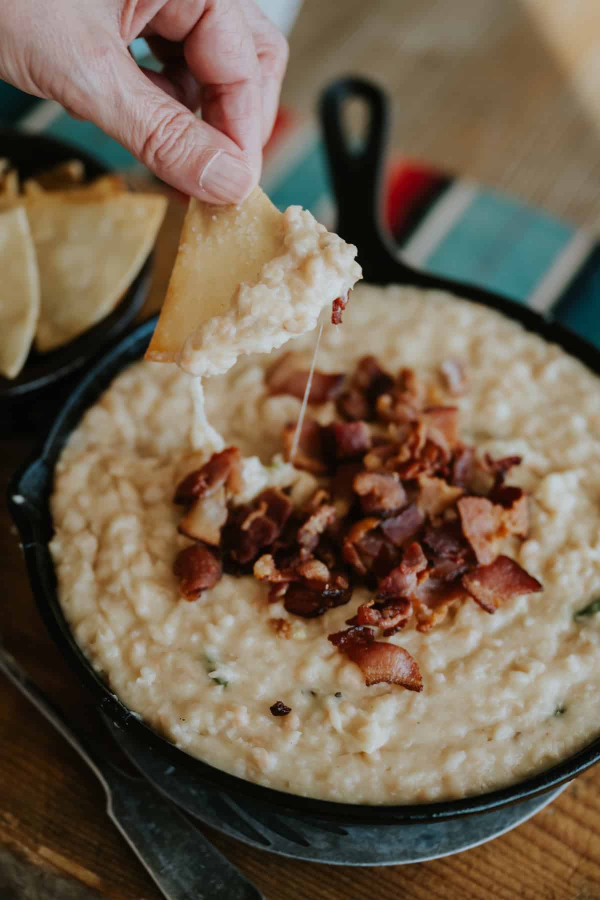 action shot of a tortilla chip being dipped into a bowl of cheesy white refried bean dip topped with extra bacon bits; the cheese is stretching from the bowl to the chip.