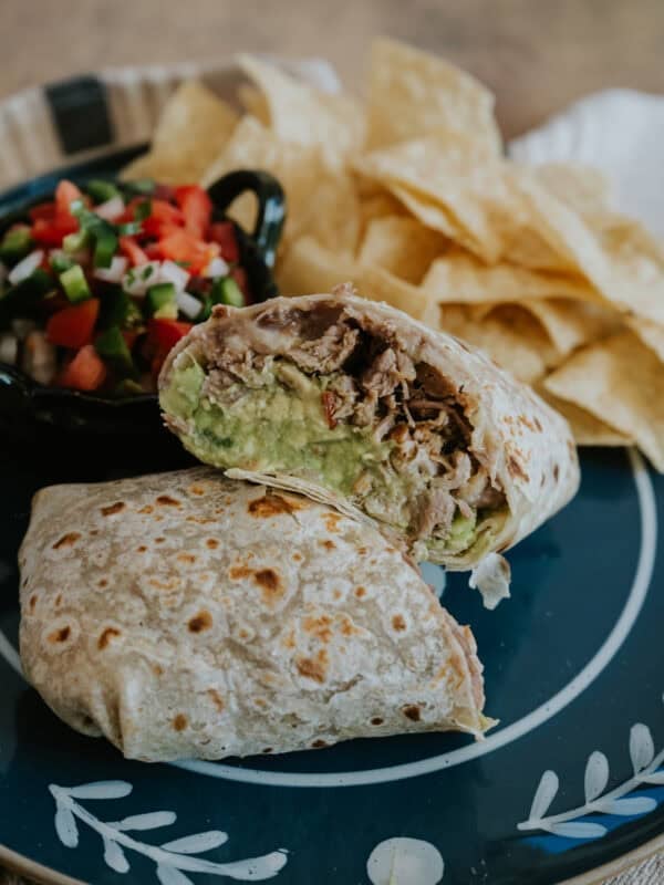 Carnitas burrito cut in half to reveal tender pulled pork, refried beans, melted cheese, and guacamole, served with tortilla chips and fresh pico de gallo on a black plate.