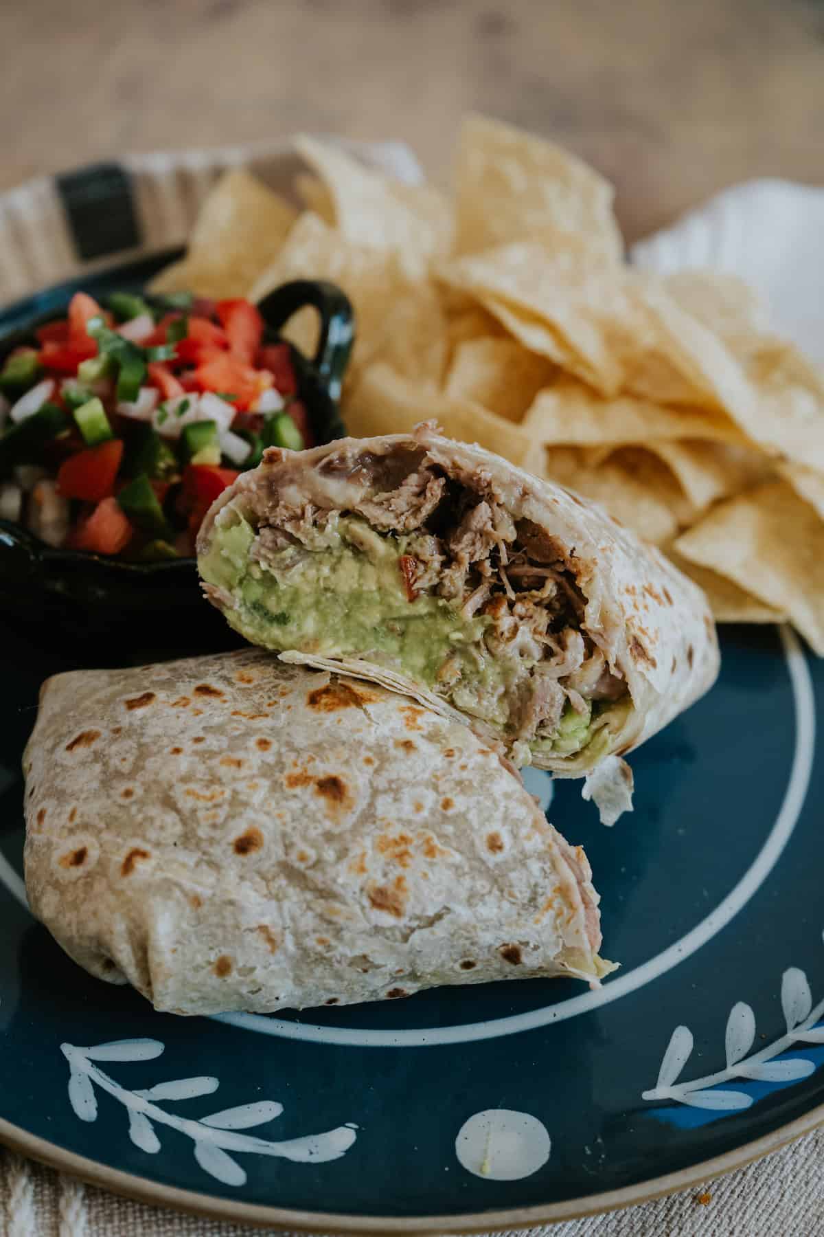 Carnitas burrito cut in half to reveal tender pulled pork, refried beans, melted cheese, and guacamole, served with tortilla chips and fresh pico de gallo on a black plate.