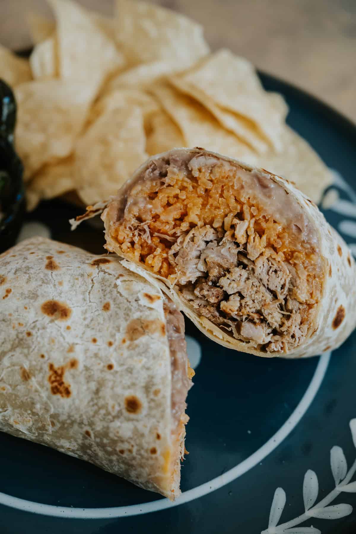Carnitas burrito sliced in half showing layers of Mexican rice, refried beans, and tender pulled pork inside a flour tortilla, served with tortilla chips on a black plate.