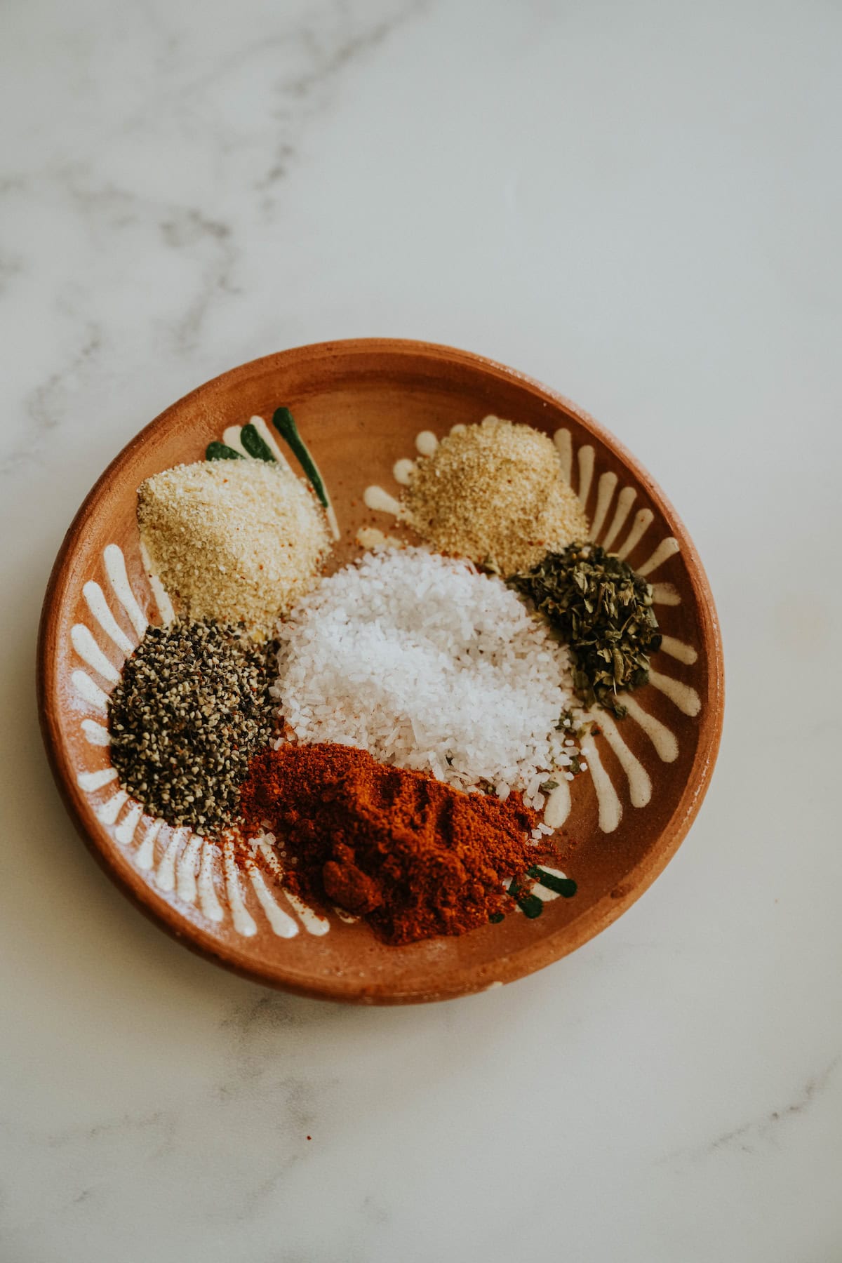 A clay plate with piles of ground spices including garlic powder, onion powder, salt, pepper, and chile powder.