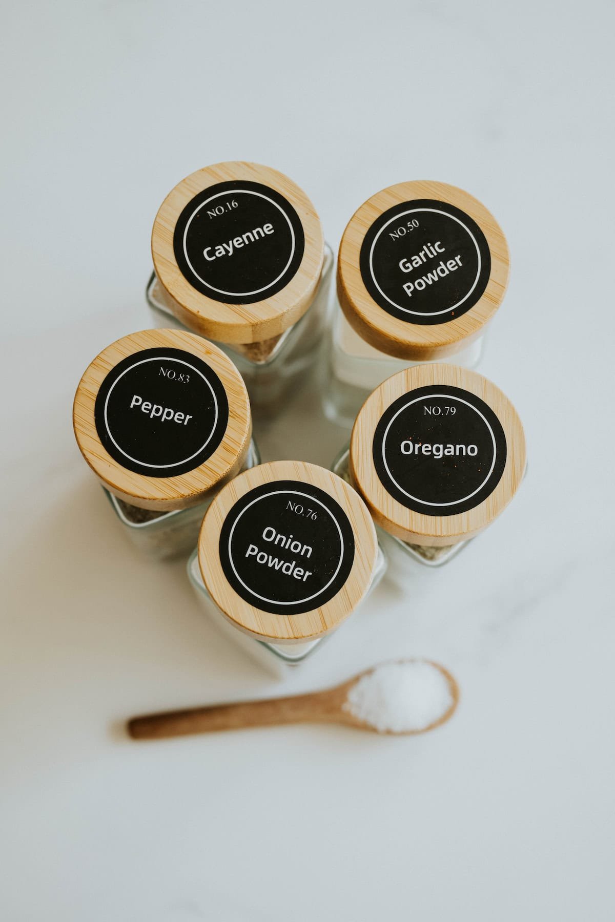 A group of spice jars labeled with seasonings stacked together with a measuring spoon on white marble.