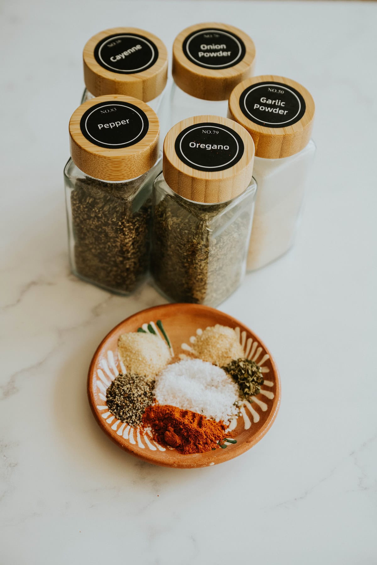 Small jars of spices labeled with black lids next to a plate of measured spices on a marble surface.