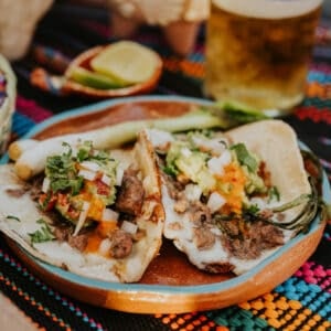 Crispy Cheese Carne Asada Tacos served on a clay plate with grilled cebollitas, salsa, and a cold chelada.