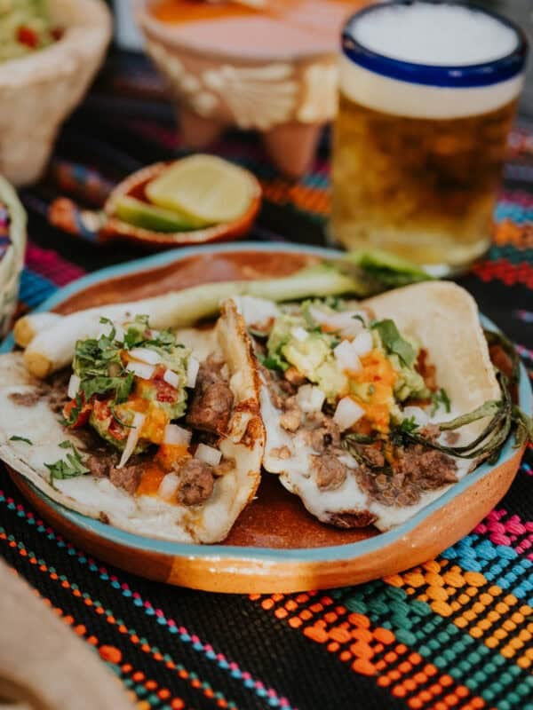 Crispy Cheese Carne Asada Tacos served on a clay plate with grilled cebollitas, salsa, and a cold chelada.