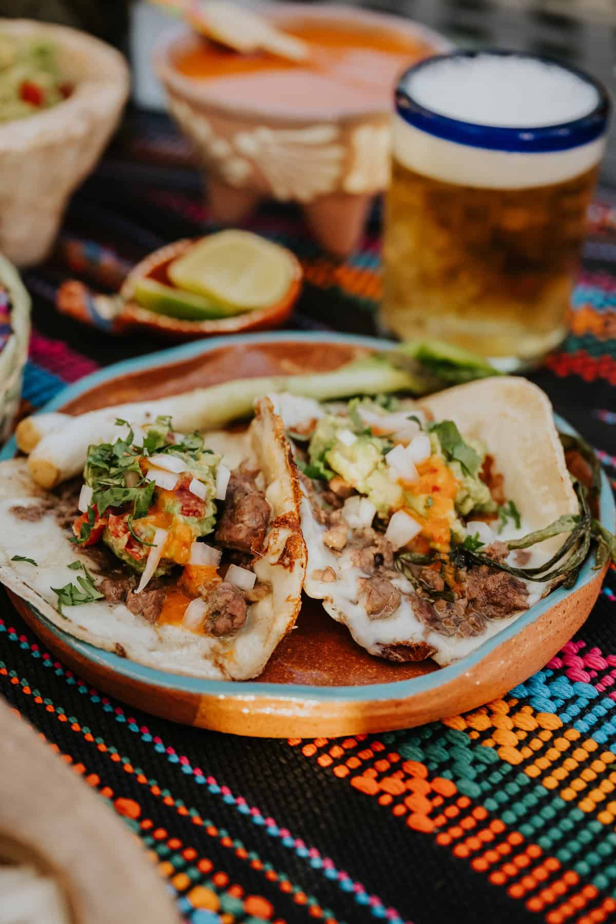 Crispy Cheese Carne Asada Tacos served on a clay plate with grilled cebollitas, salsa, and a cold chelada.