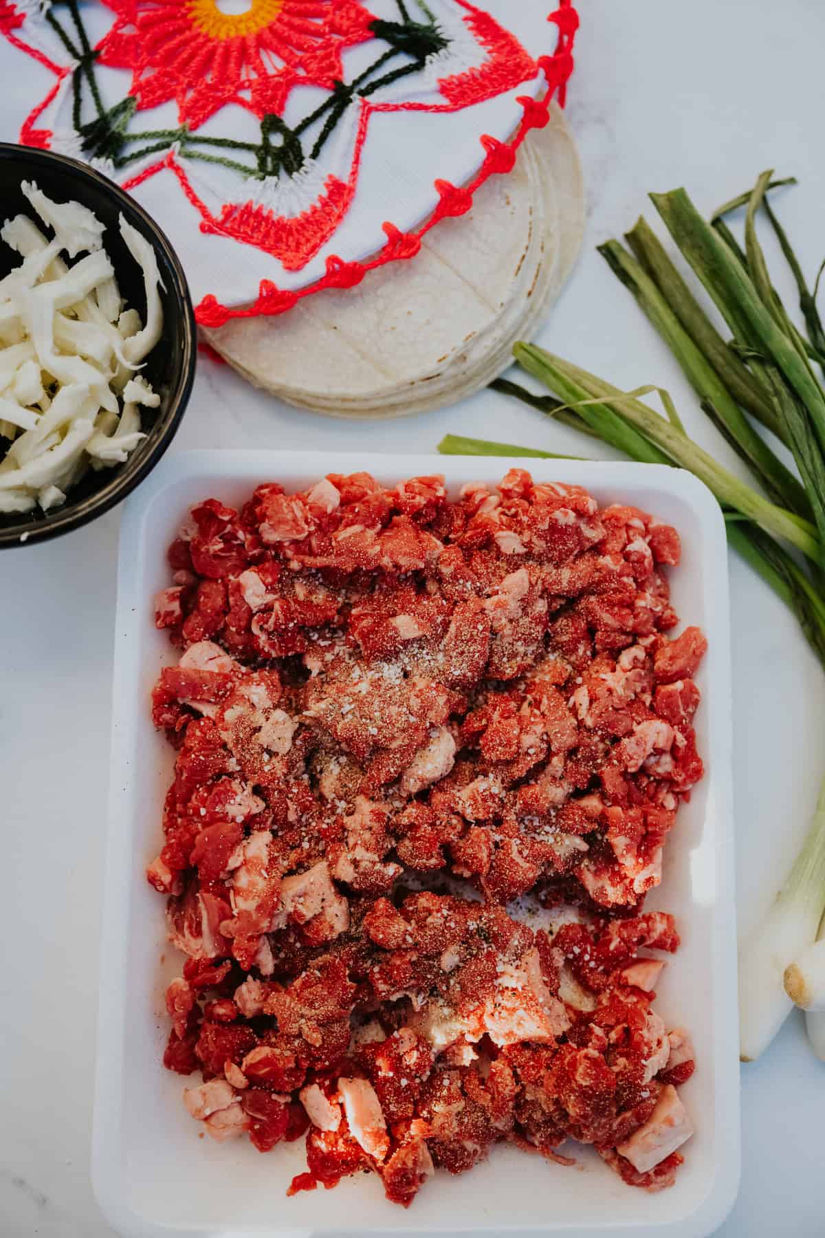 Fresh diced beef taco meat on a white plate surrounded with green onions, corn tortillas, oaxaca cheese.